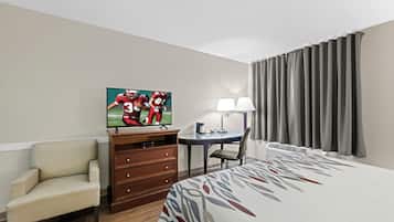 A hotel room with a bed, a TV on a wooden cabinet, a chair, a desk with a lamp, and a window with curtains.