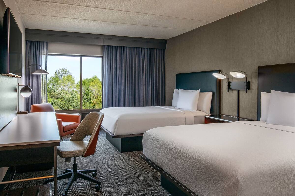 A hotel room with two beds, a desk, a chair, and a large window with curtains.
