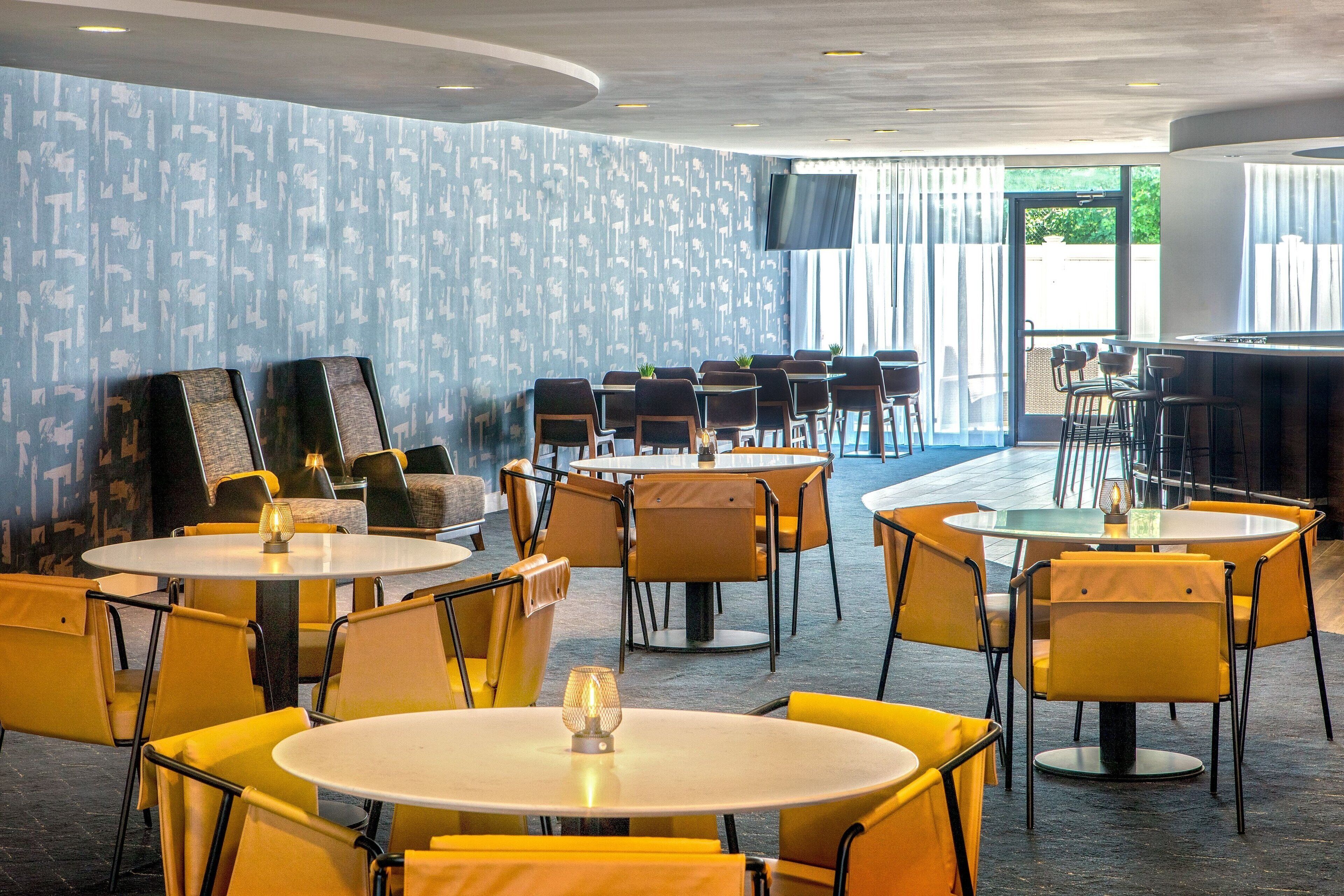 A restaurant with round tables and yellow chairs, a bar area, and a patterned wall.