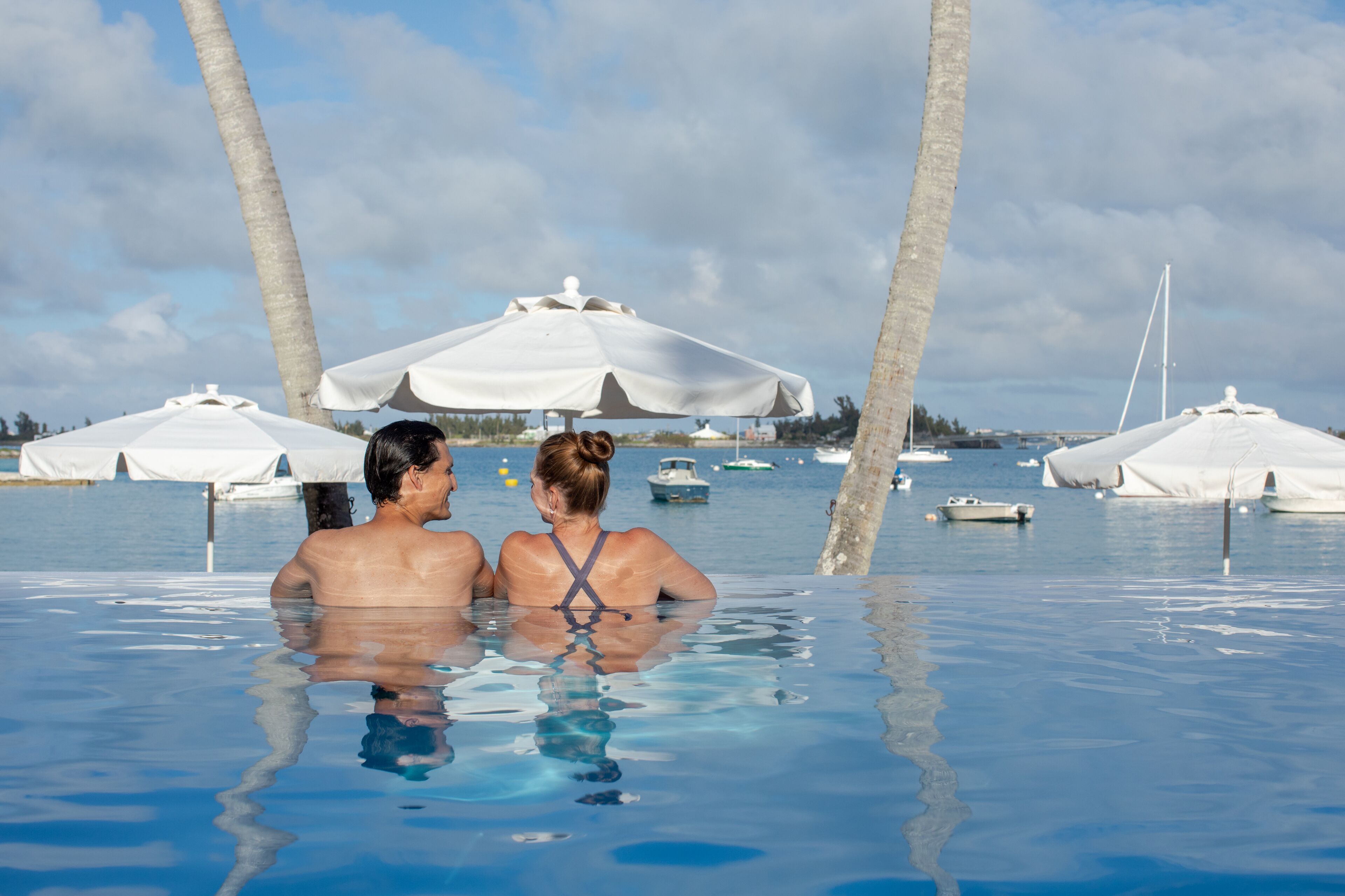 A couple in a clear infinity pool with umbrellas and palm trees in the background.