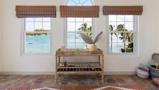 A room with three windows overlooking a beach, a wicker console table with decorative items, and a patterned rug on the floor.