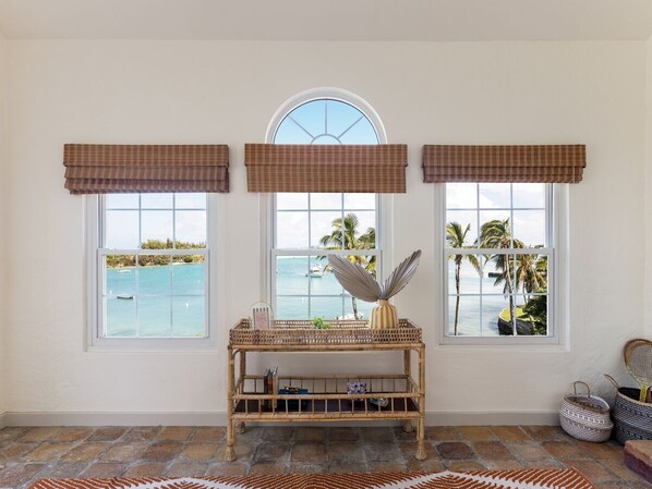 Habitación con tres ventanas que dan a una playa, una consola de mimbre con objetos decorativos y una alfombra estampada en el suelo.
