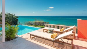 A poolside area with a view of the ocean, a lounge chair, and a small table with a bowl and a towel.