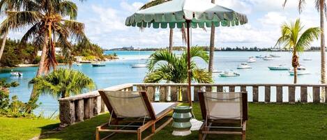 A poolside area with two lounge chairs and a table under a green umbrella, overlooking a body of water with boats and palm trees.