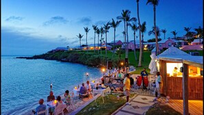 A beachfront dining area with tables and chairs, palm trees, and a wooden structure with a counter.