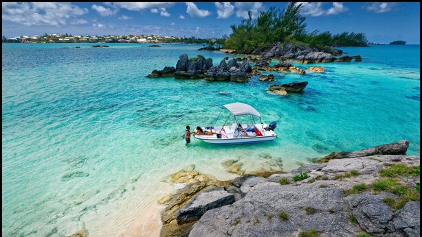 A boat anchored near a rocky shoreline with clear turquoise water and a distant view of a coastal town.