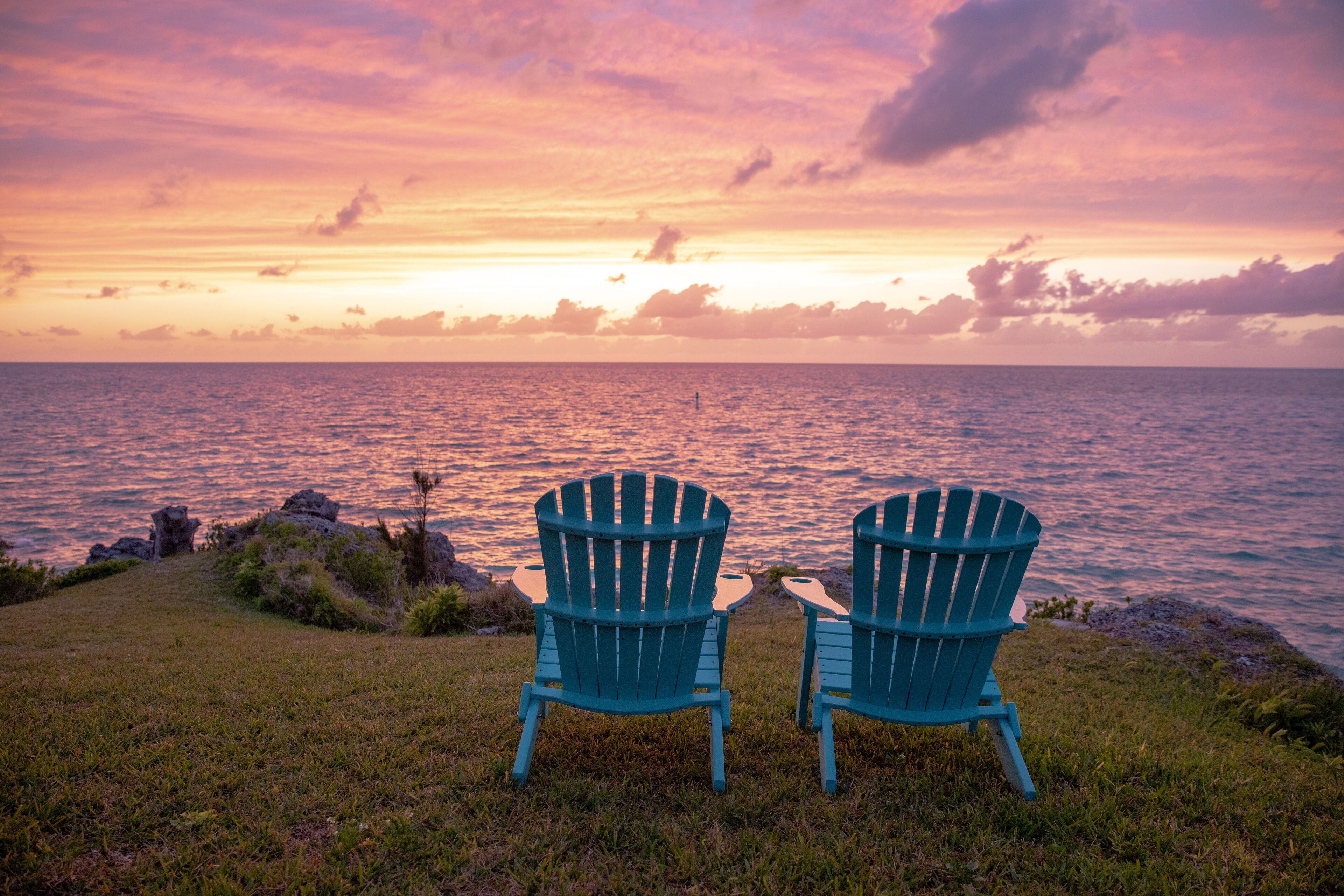 Two Adirondack chairs facing a sunset over the ocean.