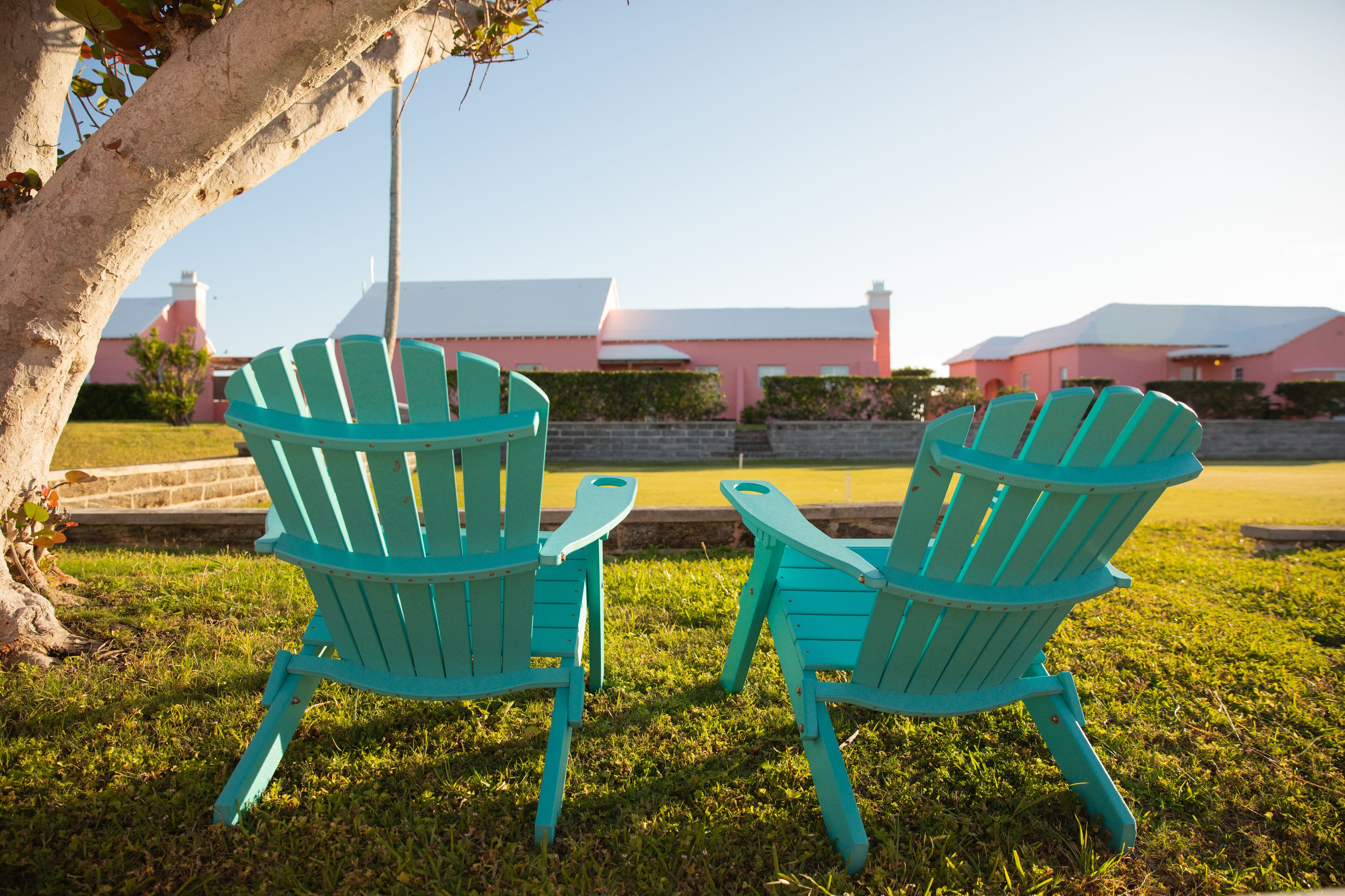 Two turquoise Adirondack chairs facing each other on a grassy lawn with a pink building in the background.