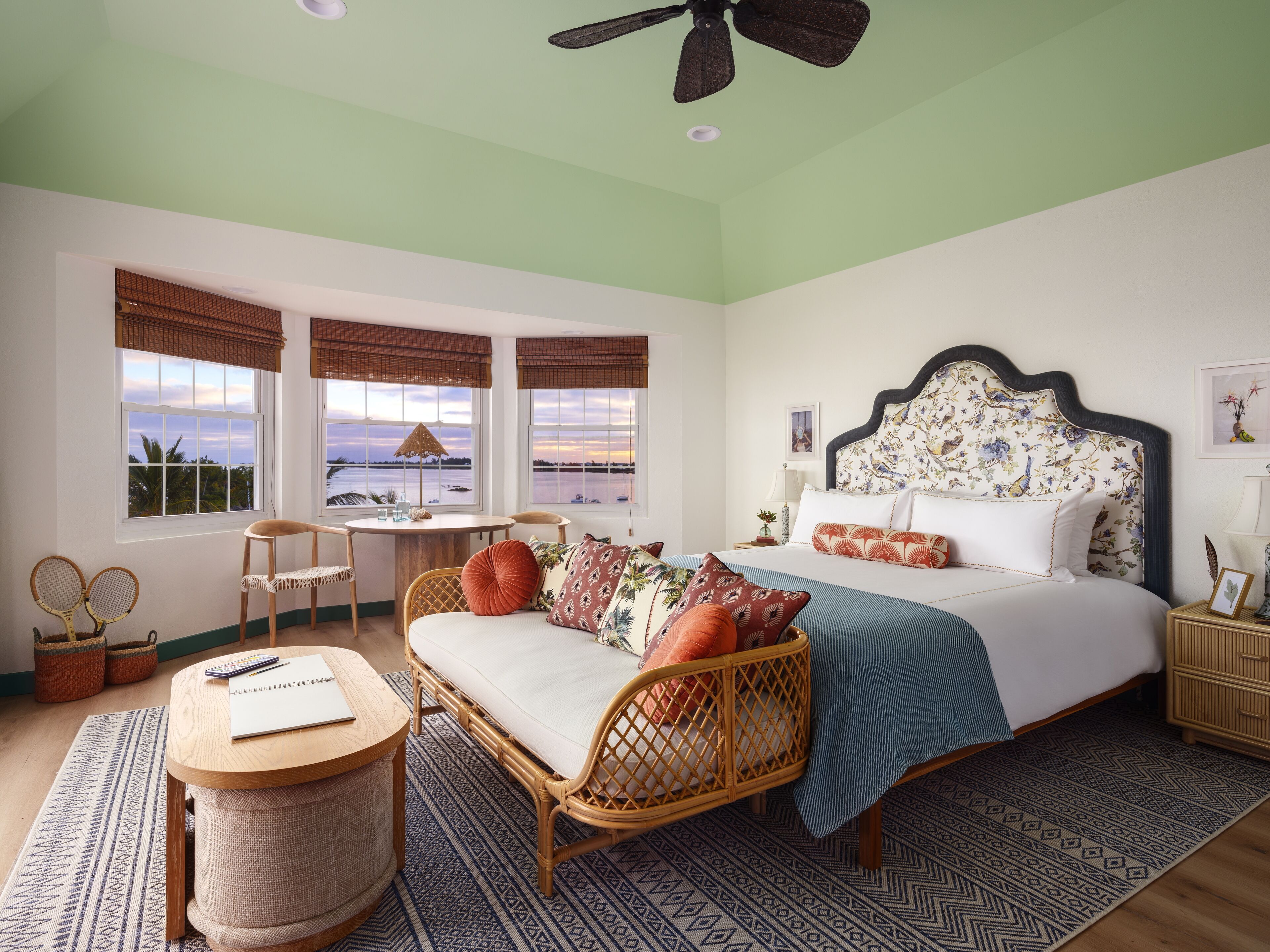 Un dormitorio con cama, sofá, una mesa redonda y una silla. Hay ventanas con cortinas, un ventilador de techo y vista al mar.