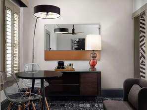 A modern hotel room with a round dining table, a glass chair, a dark wooden cabinet, a decorative lamp, and a large mirror on the wall.