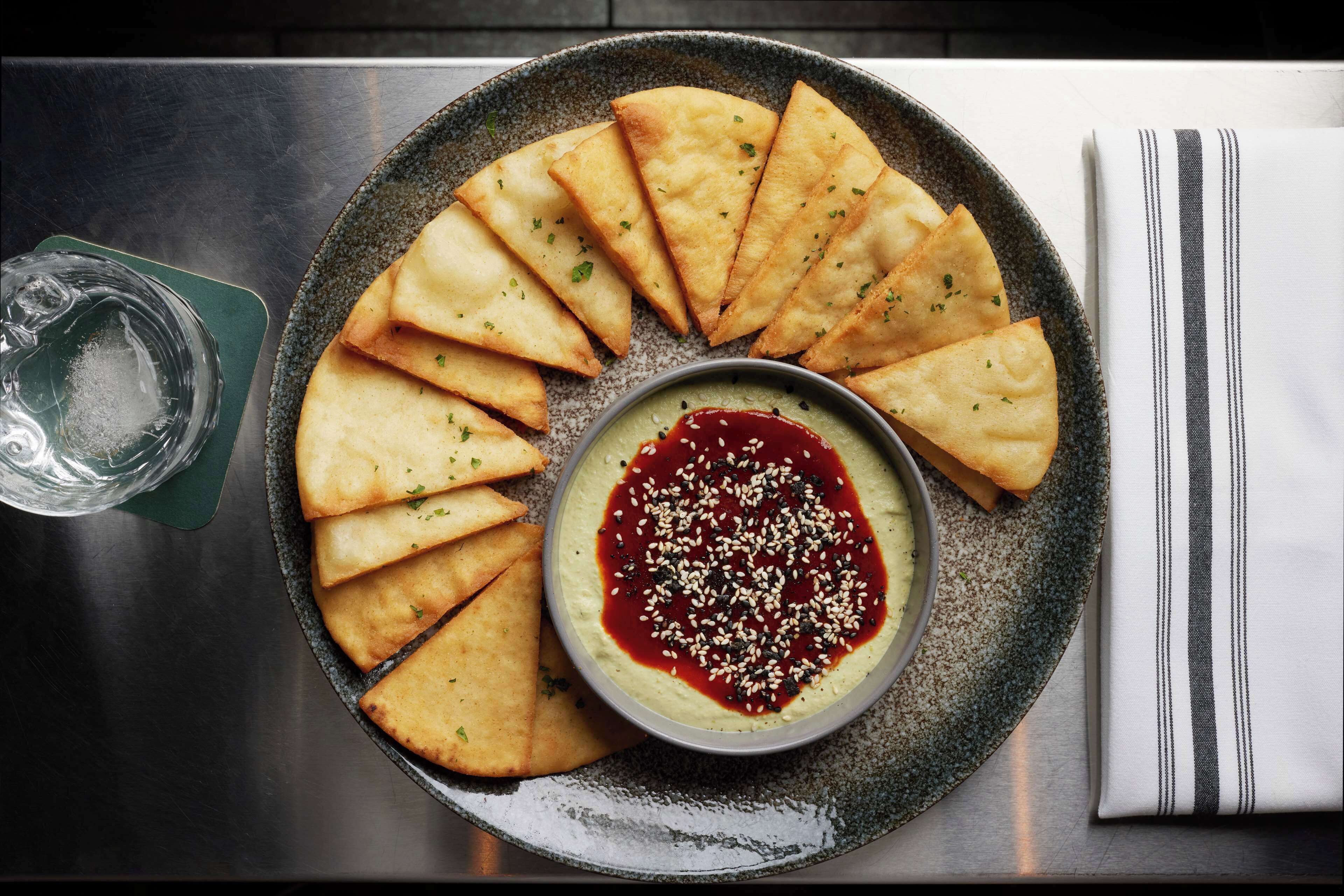 A plate of triangular pastries with a dipping sauce, a glass of water, and a striped napkin on a table.