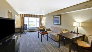 A hotel room with a flat-screen TV, a sofa, a coffee table, a desk with a lamp, and a view of the outdoors. - Hampton Inn Mobile-East Bay/Daphne (Daphne)