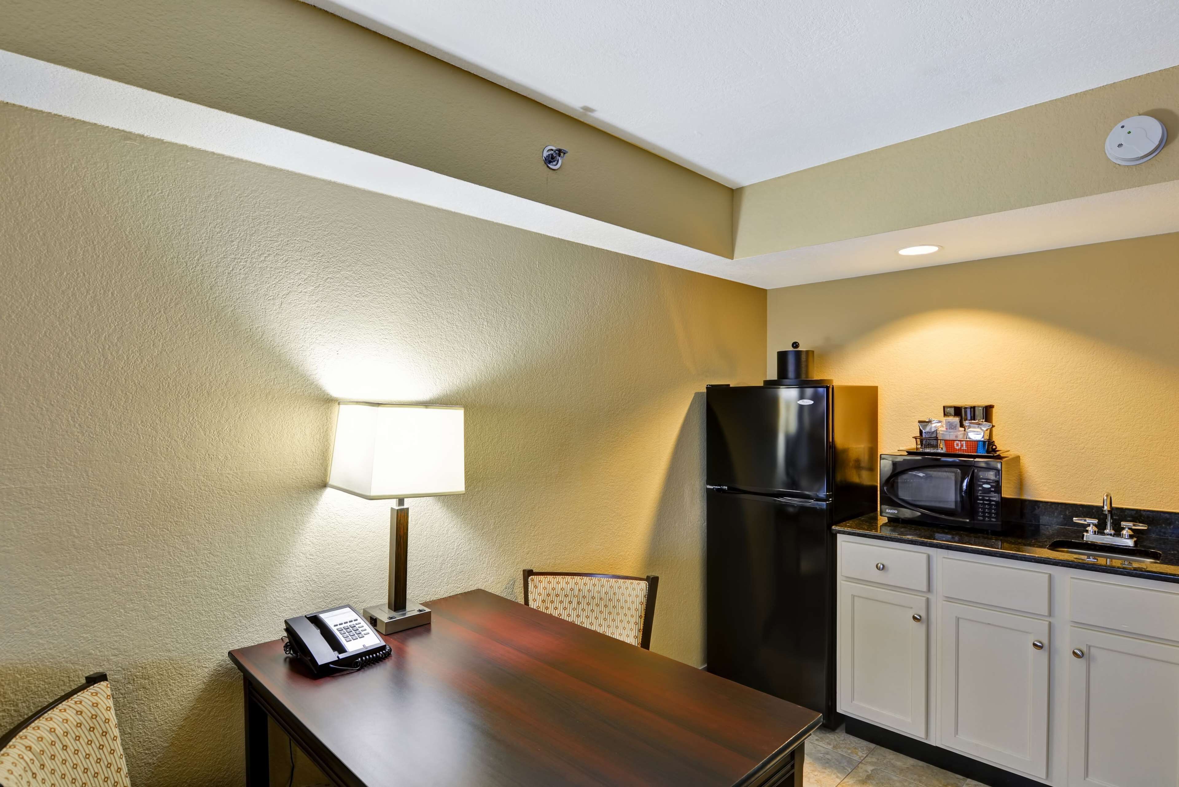 A compact kitchen with a refrigerator, microwave, and a small countertop. There is a table with a lamp and a telephone.