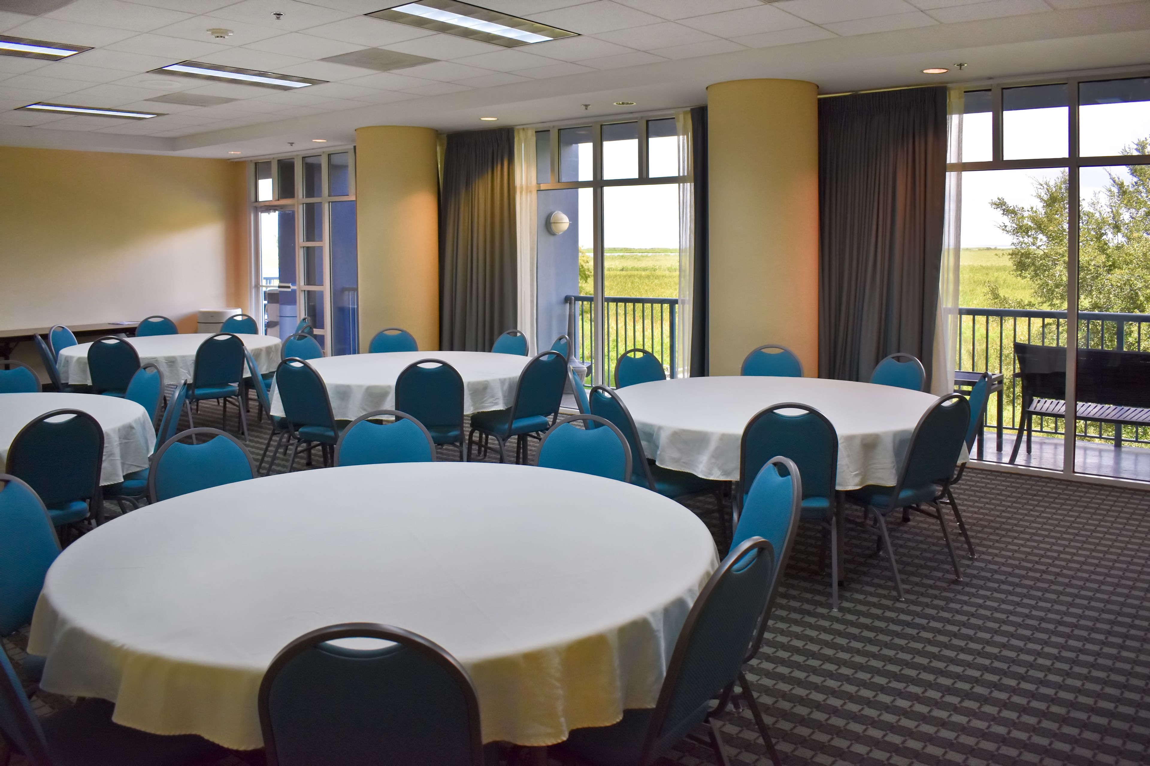 Un salón de conferencias con varias mesas redondas y sillas azules, ventanas amplias con cortinas y vistas al exterior.