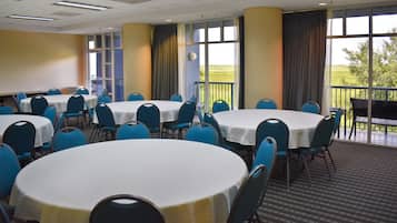 Un salón de conferencias con varias mesas redondas y sillas azules, ventanas amplias con cortinas y vistas al exterior.