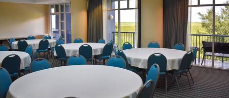 Une salle de conférence dotée de plusieurs tables rondes et de chaises bleues, de grandes fenêtres avec des rideaux et offrant une vue sur l’extérieur.