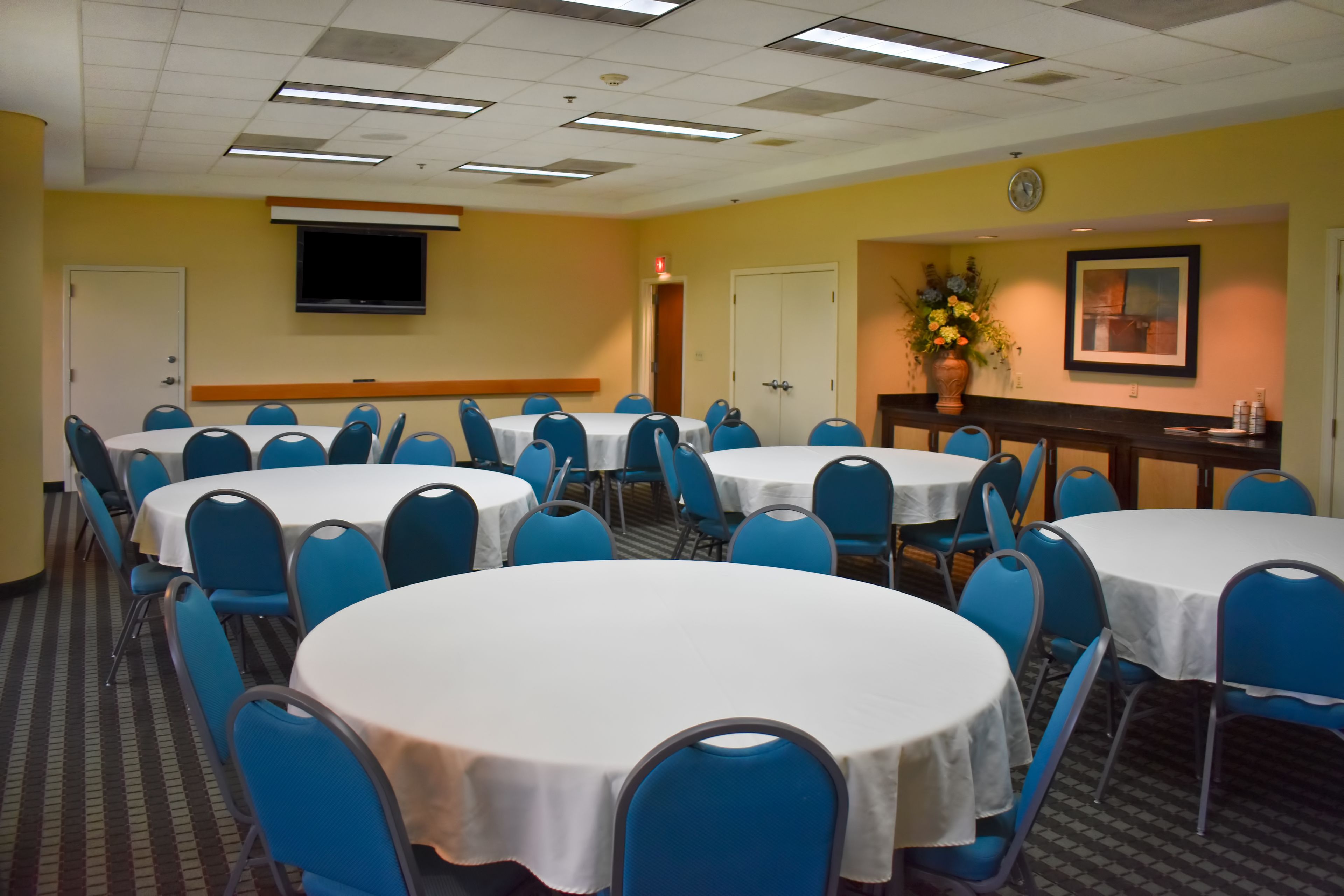 A conference room with multiple round tables and blue chairs, a clock on the wall, and a television mounted above a reception desk.