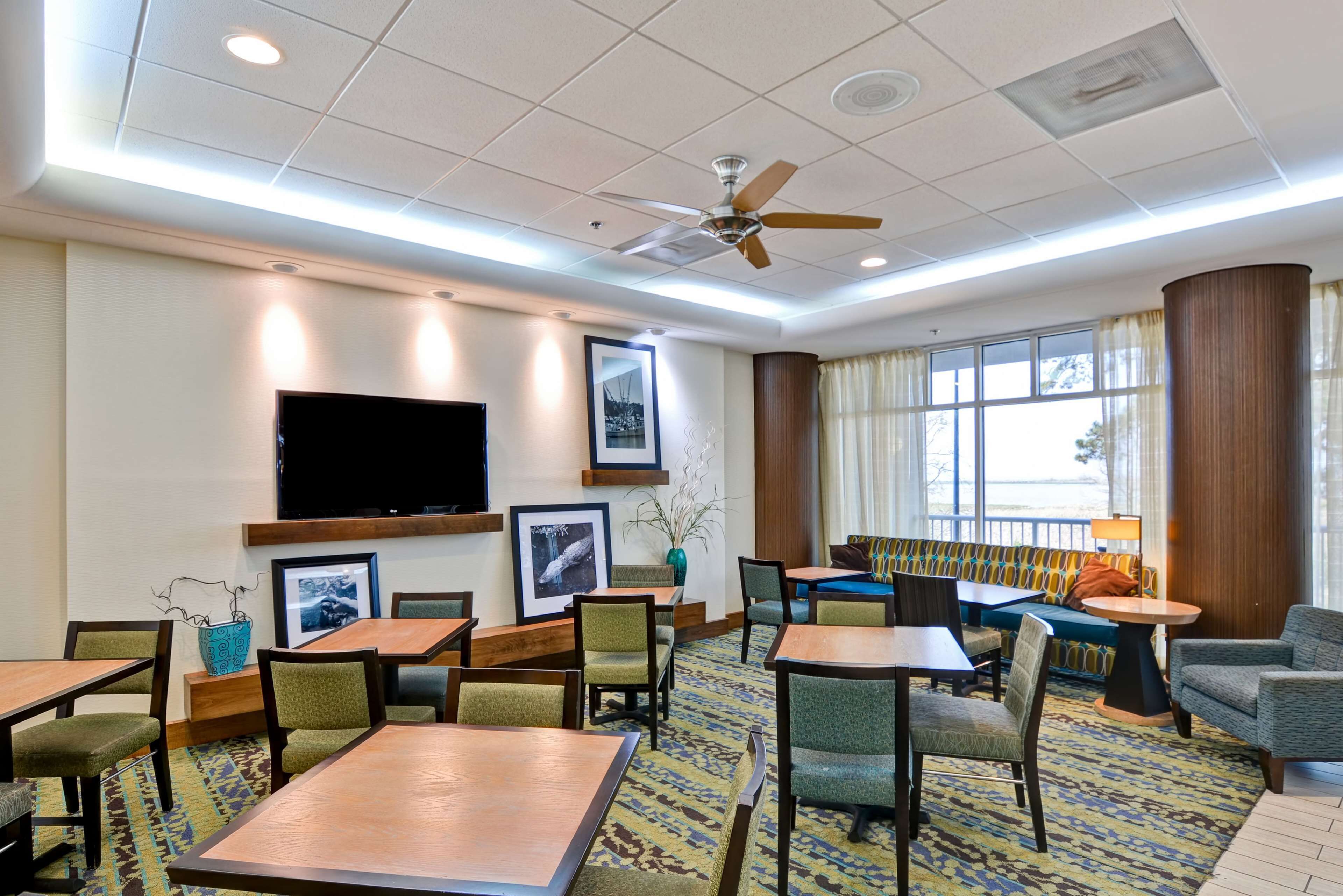 A hotel lobby with a large flat-screen TV, multiple seating areas including a sofa and chairs, a ceiling fan, and a large window with curtains.
