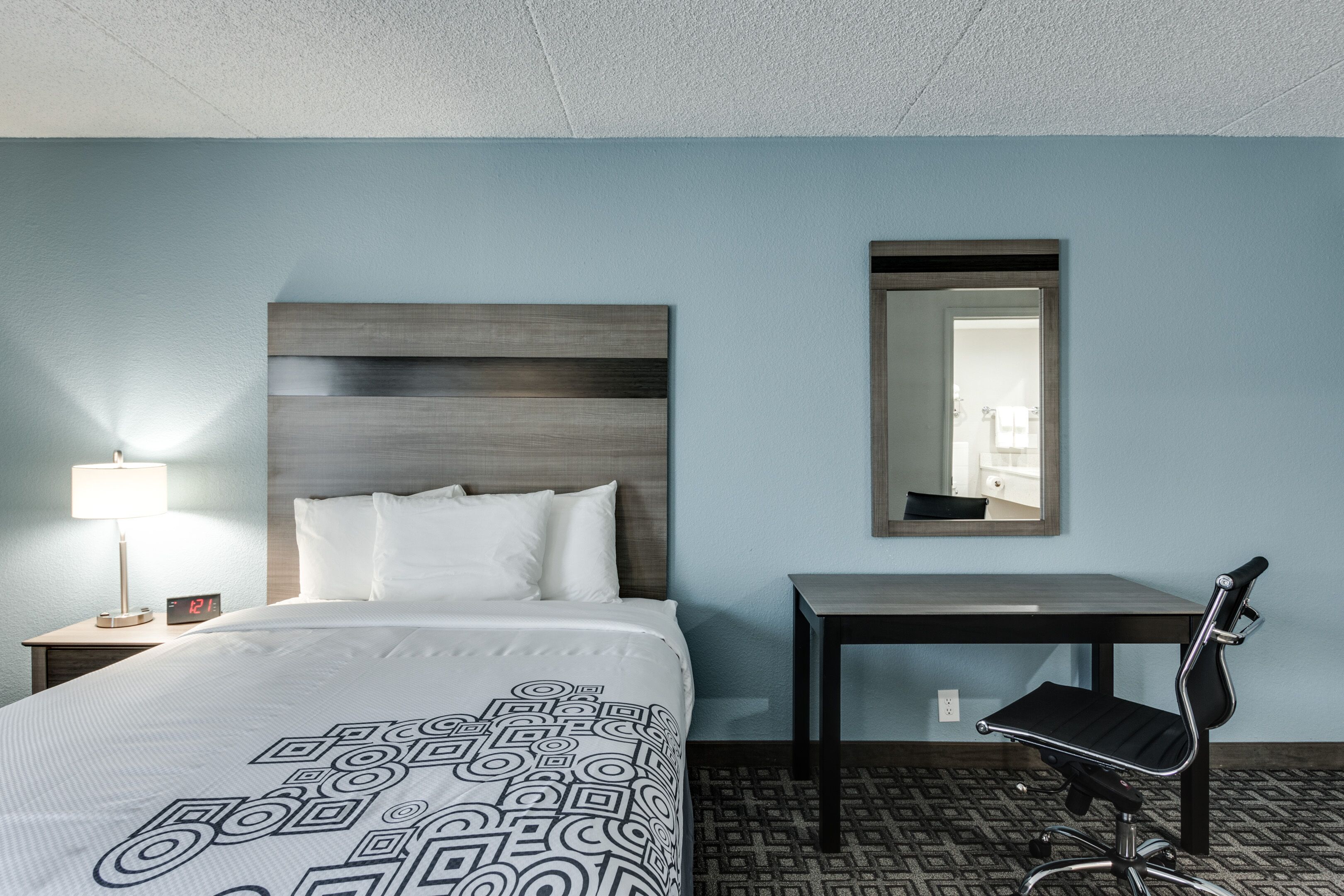 Una habitación de hotel con cama, escritorio, silla, mesita de noche con lámpara y espejo en la pared.