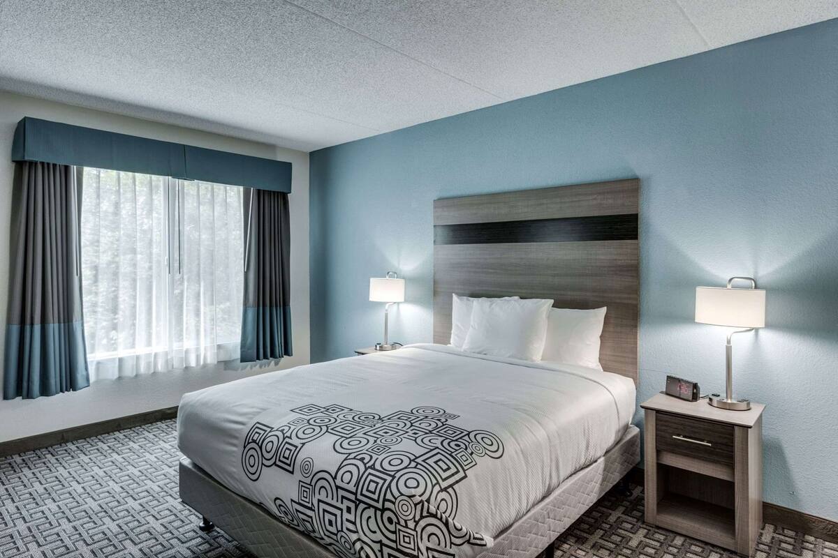 A hotel room with a large bed, two bedside tables with lamps, a headboard, and a window with curtains.