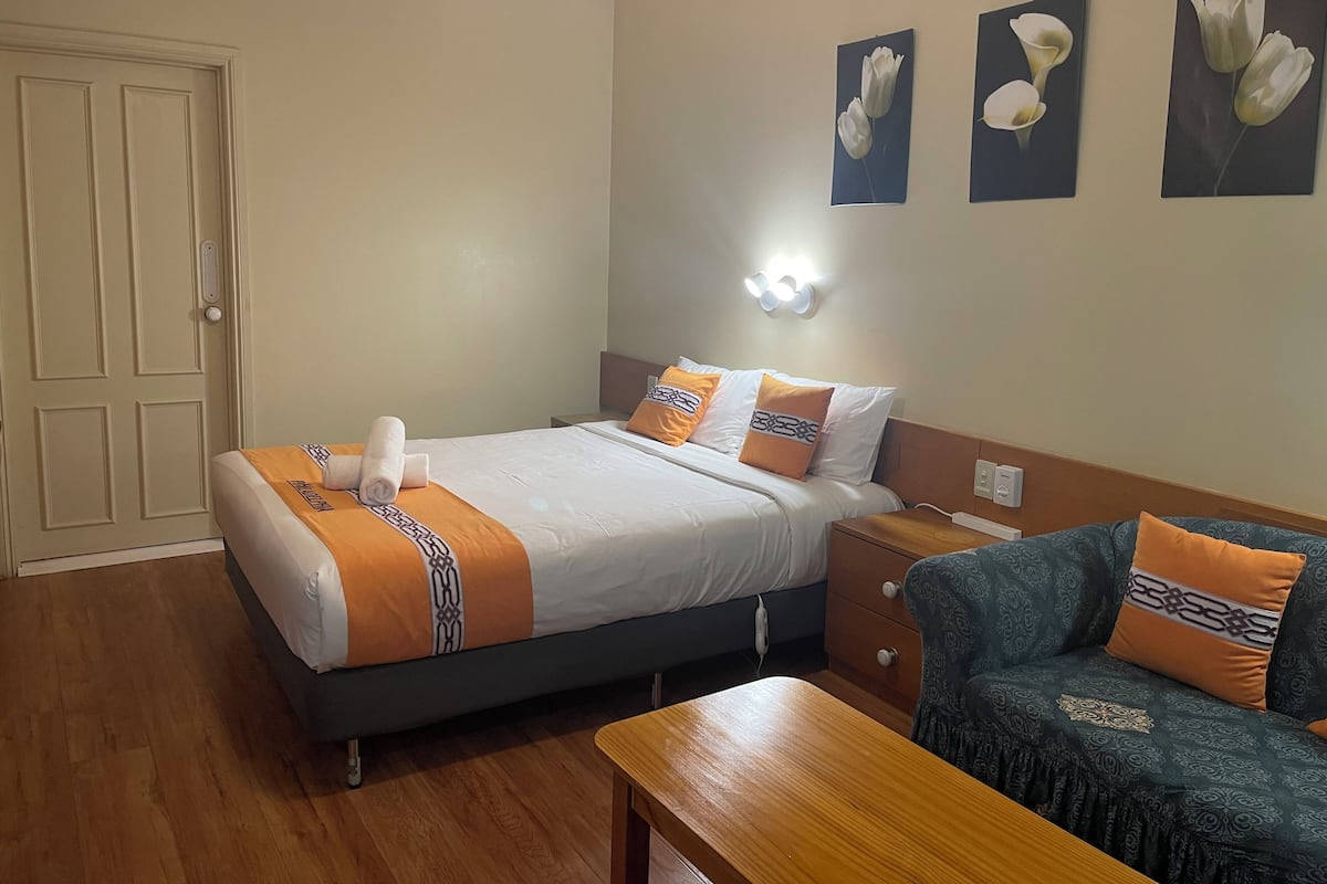 A hotel room with a bed, a nightstand, a wooden table, and a sofa. There are three wall-mounted light fixtures above the bed.