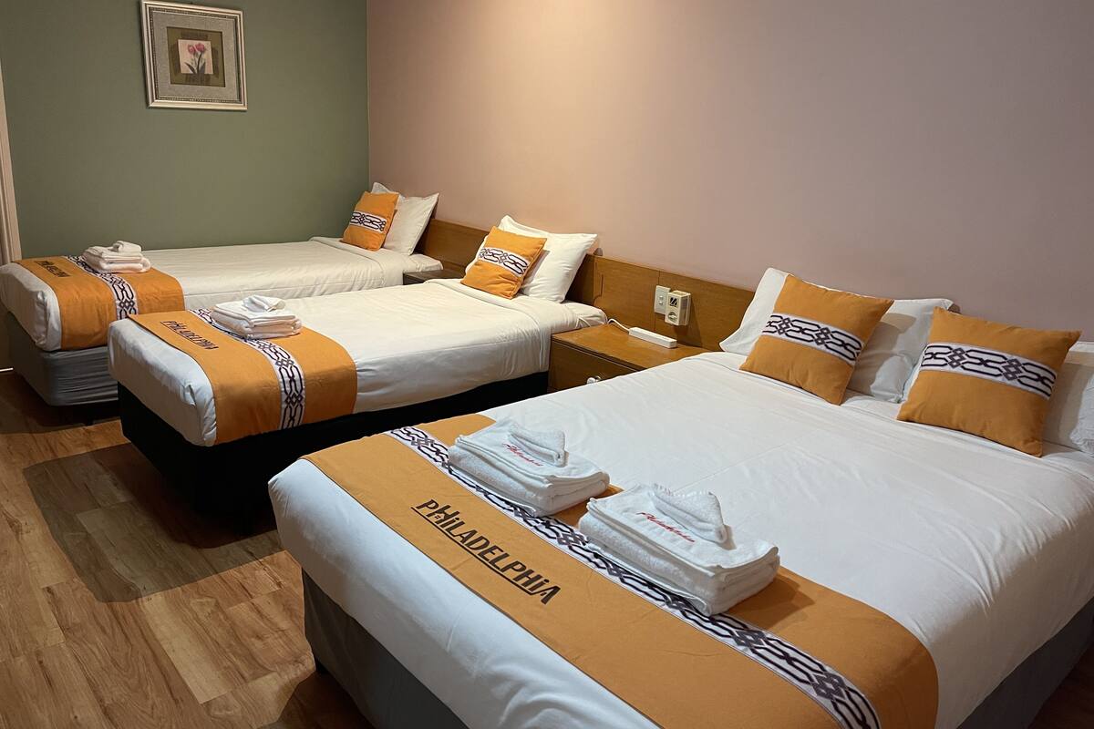 A hotel room with three beds, each with a white and orange bedspread and matching pillows. There is a framed picture on the wall and a bedside table with a lamp.