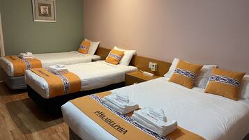 A hotel room with three beds, each with a white and orange bedspread and matching pillows. There is a framed picture on the wall and a bedside table with a lamp.