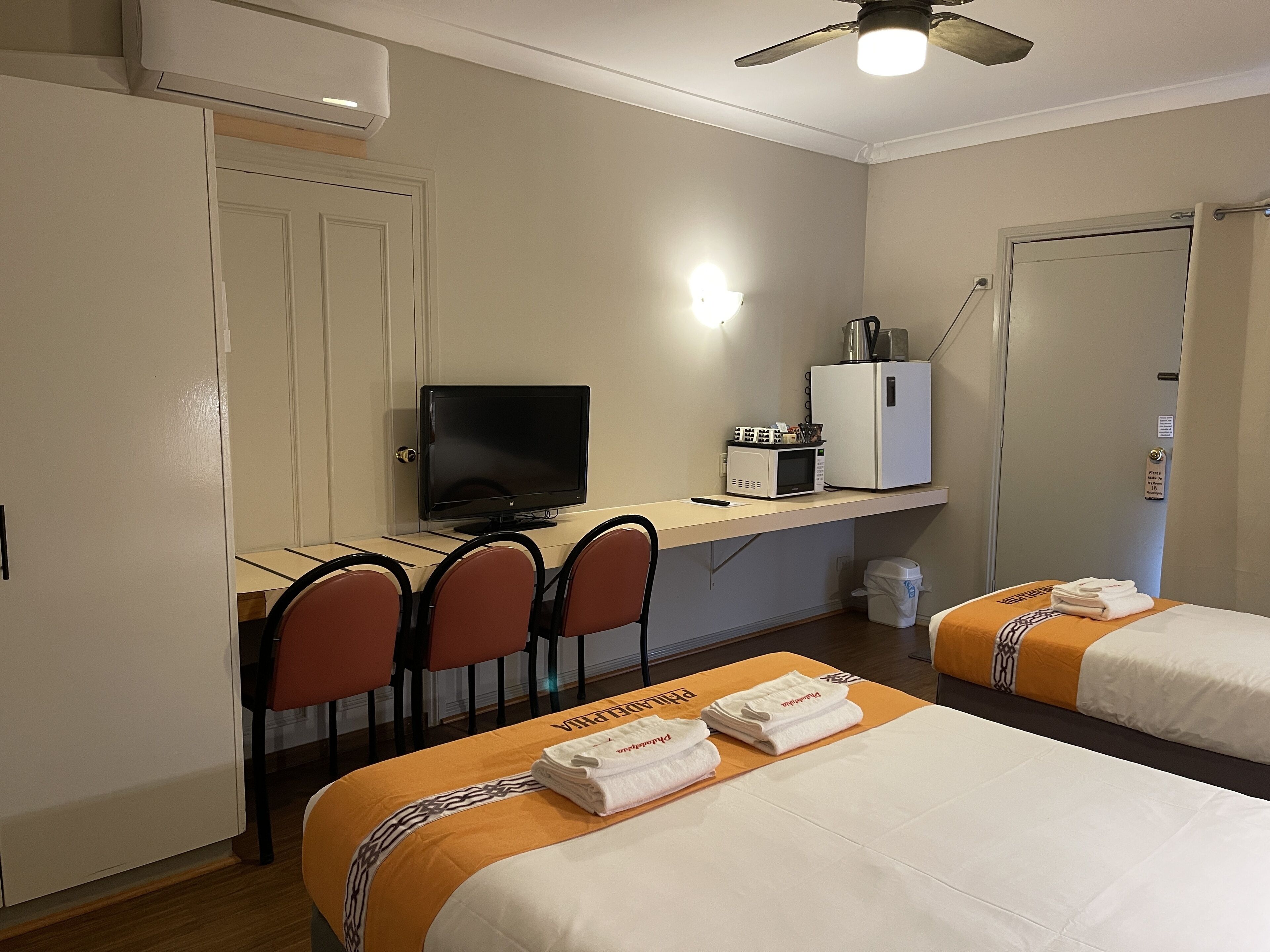A hotel room with a desk, a television, a refrigerator, two beds with towels, and a ceiling fan.