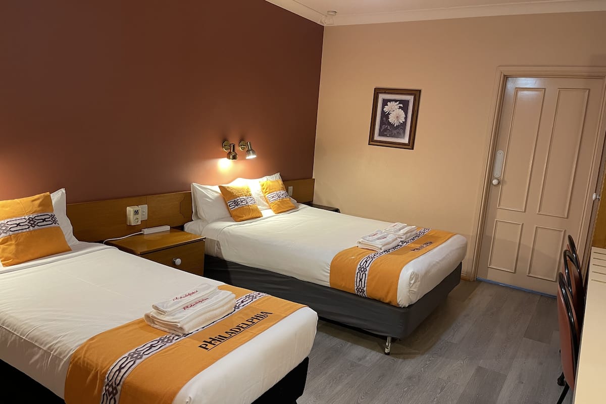 A hotel room with two beds, each with a Philadelphia-themed bedspread and matching pillows. There are bedside tables with lamps, a framed picture on the wall, and a door to the right.