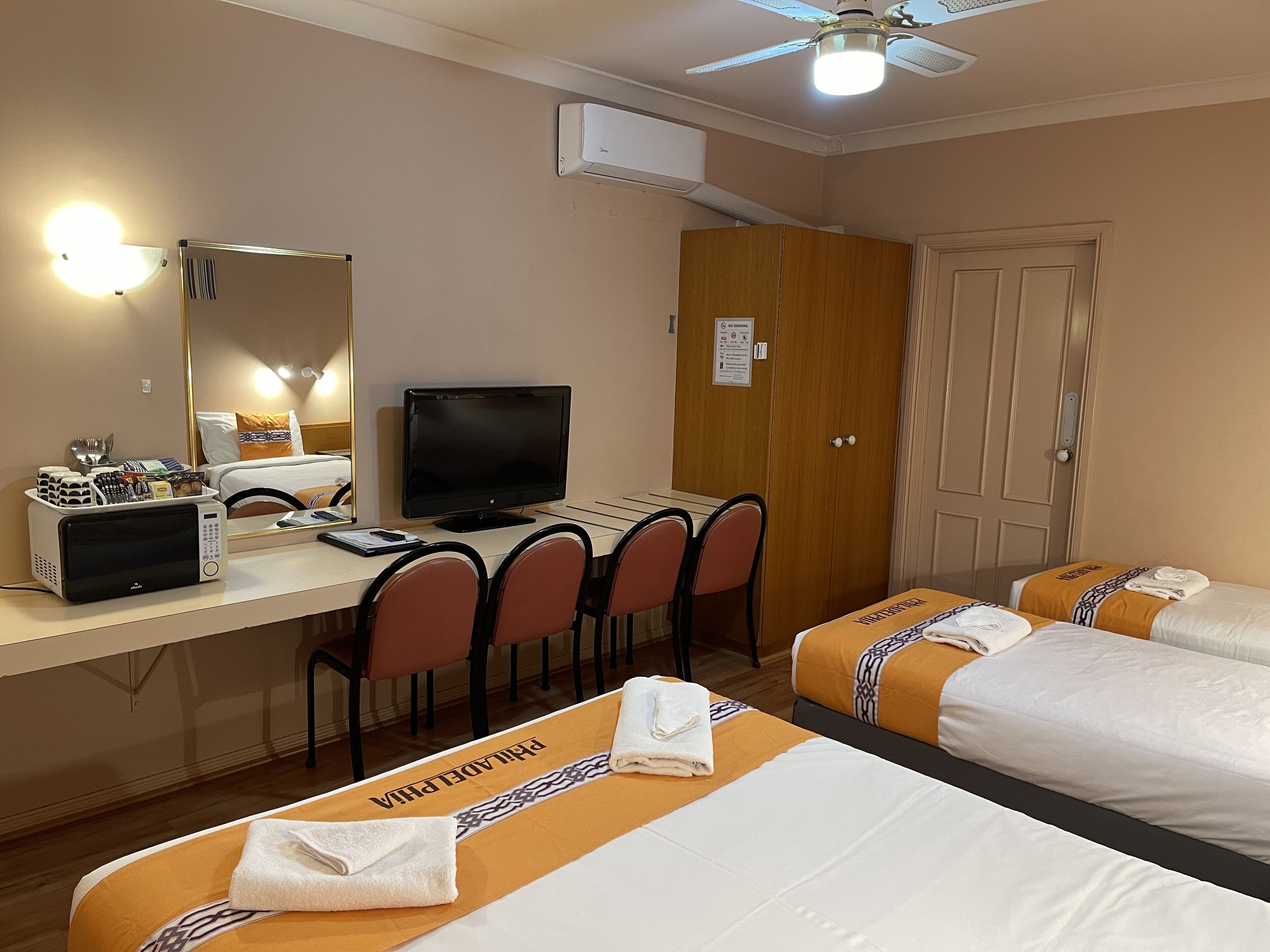 Camera d'albergo con scrivania, due letti, televisore, forno a microonde e ventilatore a soffitto.