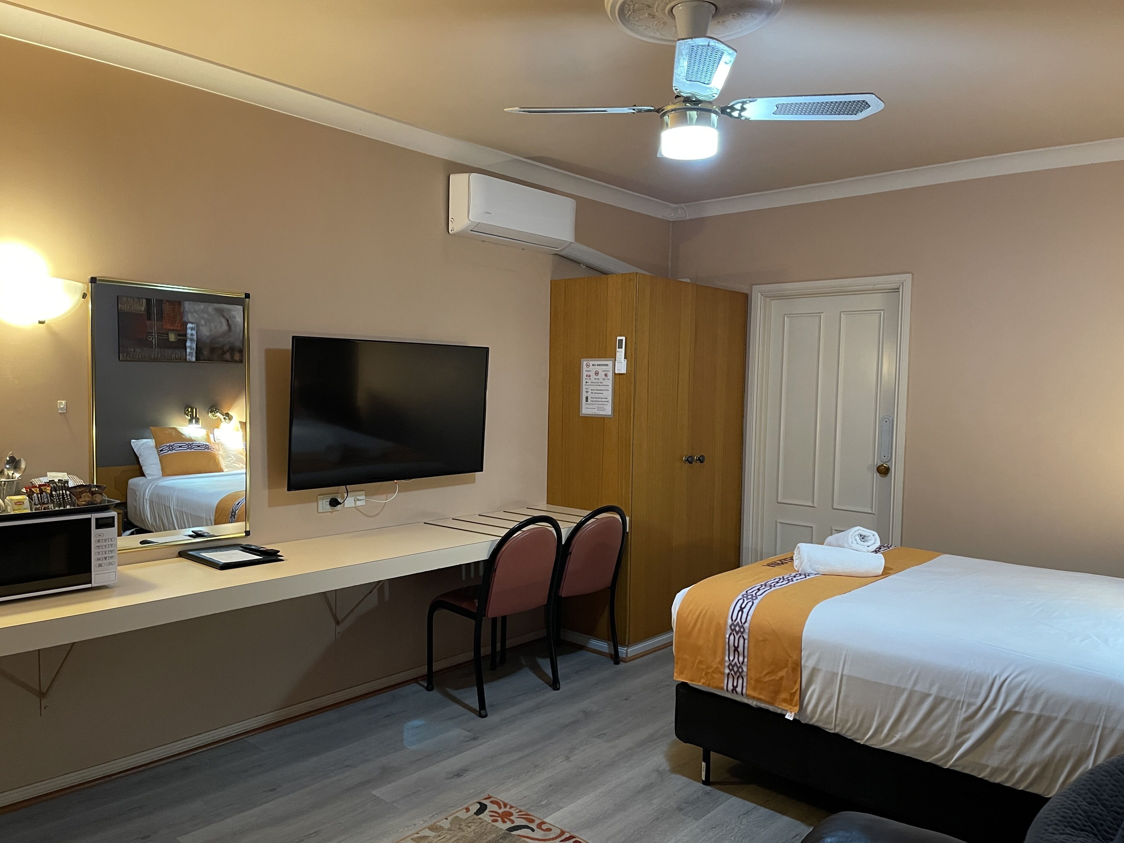 Ein Hotelzimmer mit Bett, Schreibtisch, Stuhl, Fernseher und Deckenventilator.