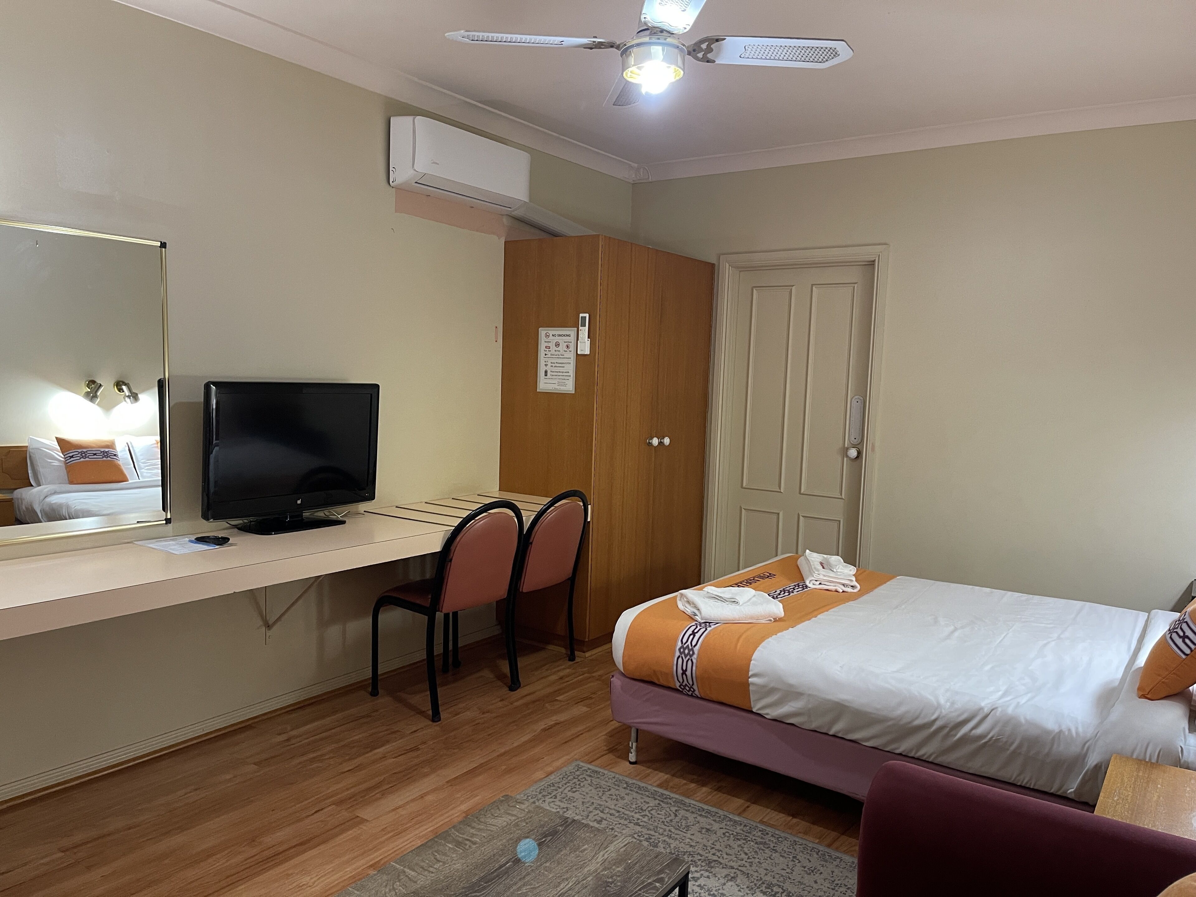 A hotel room with a bed, a desk with a computer, a chair, a ceiling fan, and an air conditioning unit.