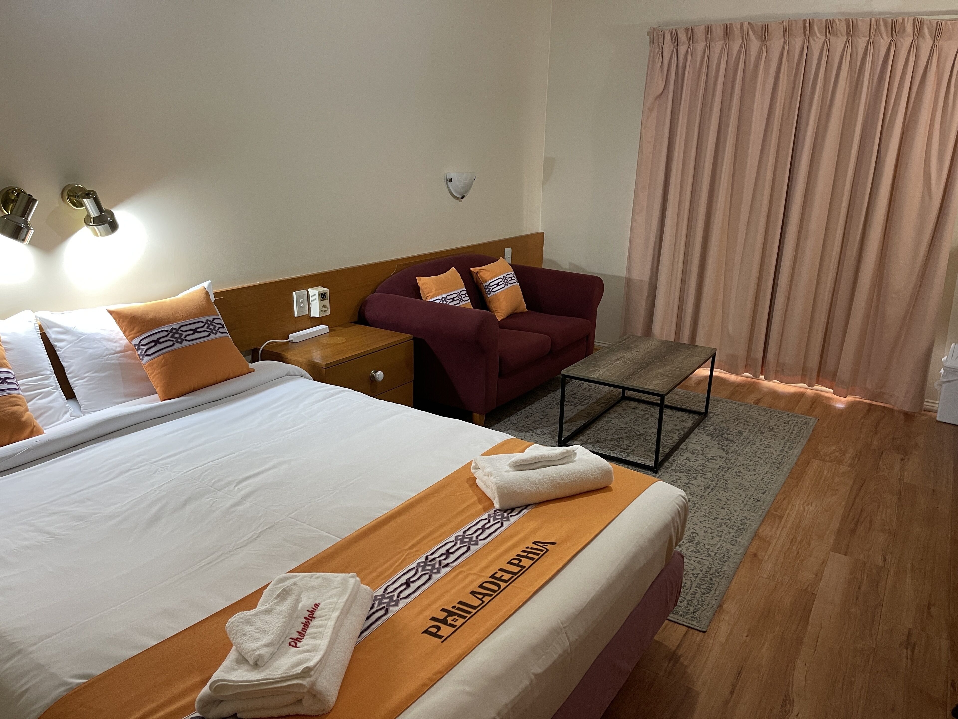 A hotel room with a bed, a nightstand, a chair, a table, and a window with curtains.