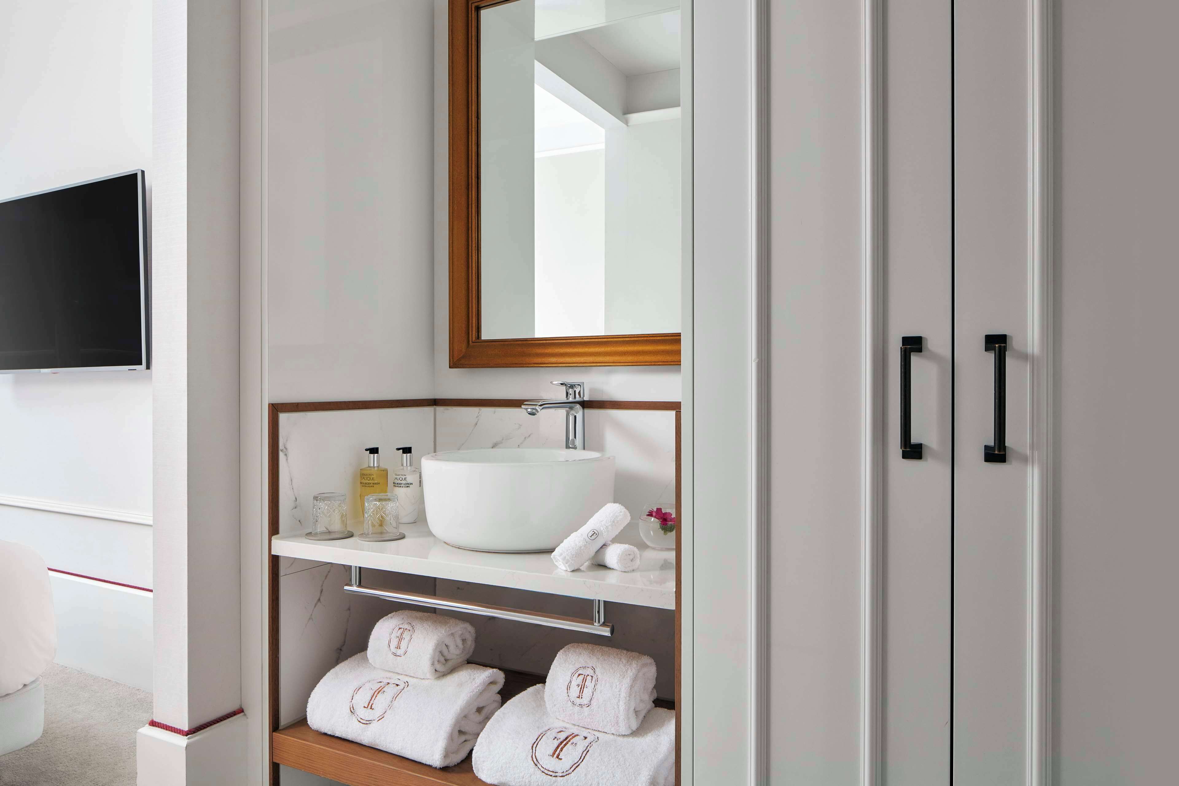 A bathroom with a large mirror, a white sink, a shelf with toiletries, and a towel rack with towels.