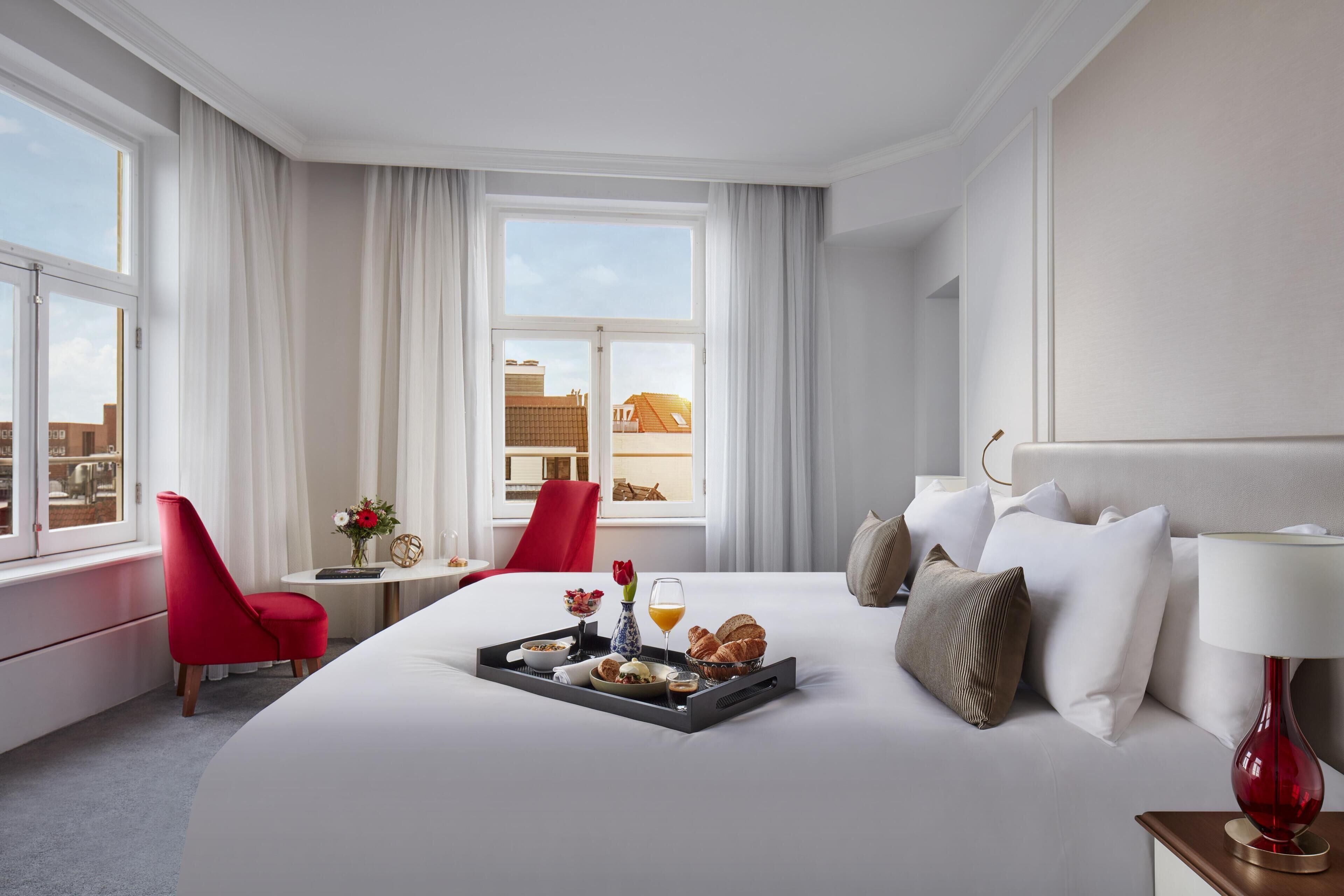 A hotel room with a large bed, a red chair, a small table with a tray of breakfast, and a view of buildings outside.