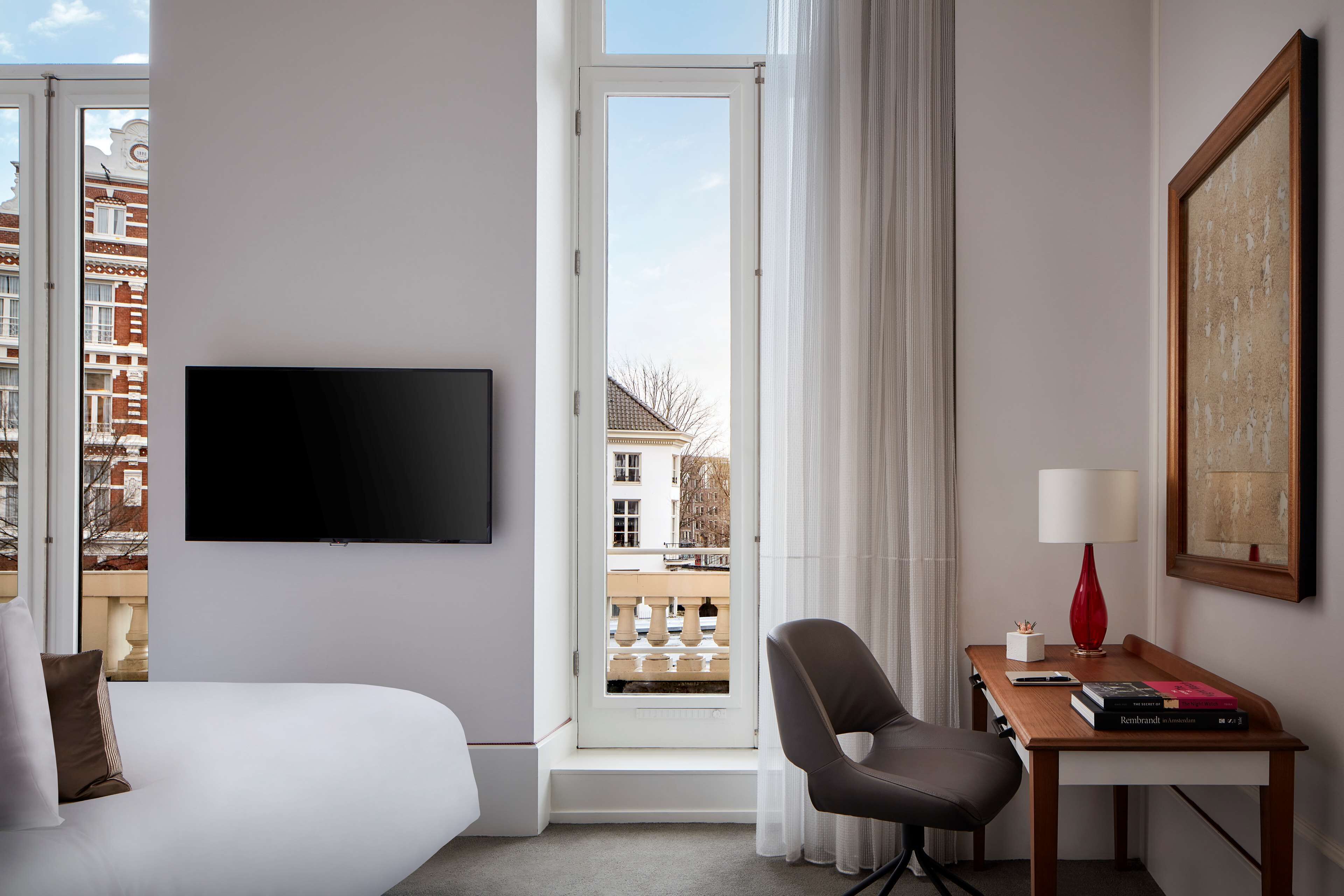 A hotel room with a bed, a desk, a chair, a television, a window with a view of buildings, and a lamp.