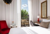 A hotel room with a bed, a red chair, a small table with a vase of flowers, a desk with a lamp, and a view of the outside.