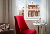 A room with a red chair, a small round table with a book and a vase, and a window with a view of a building.
