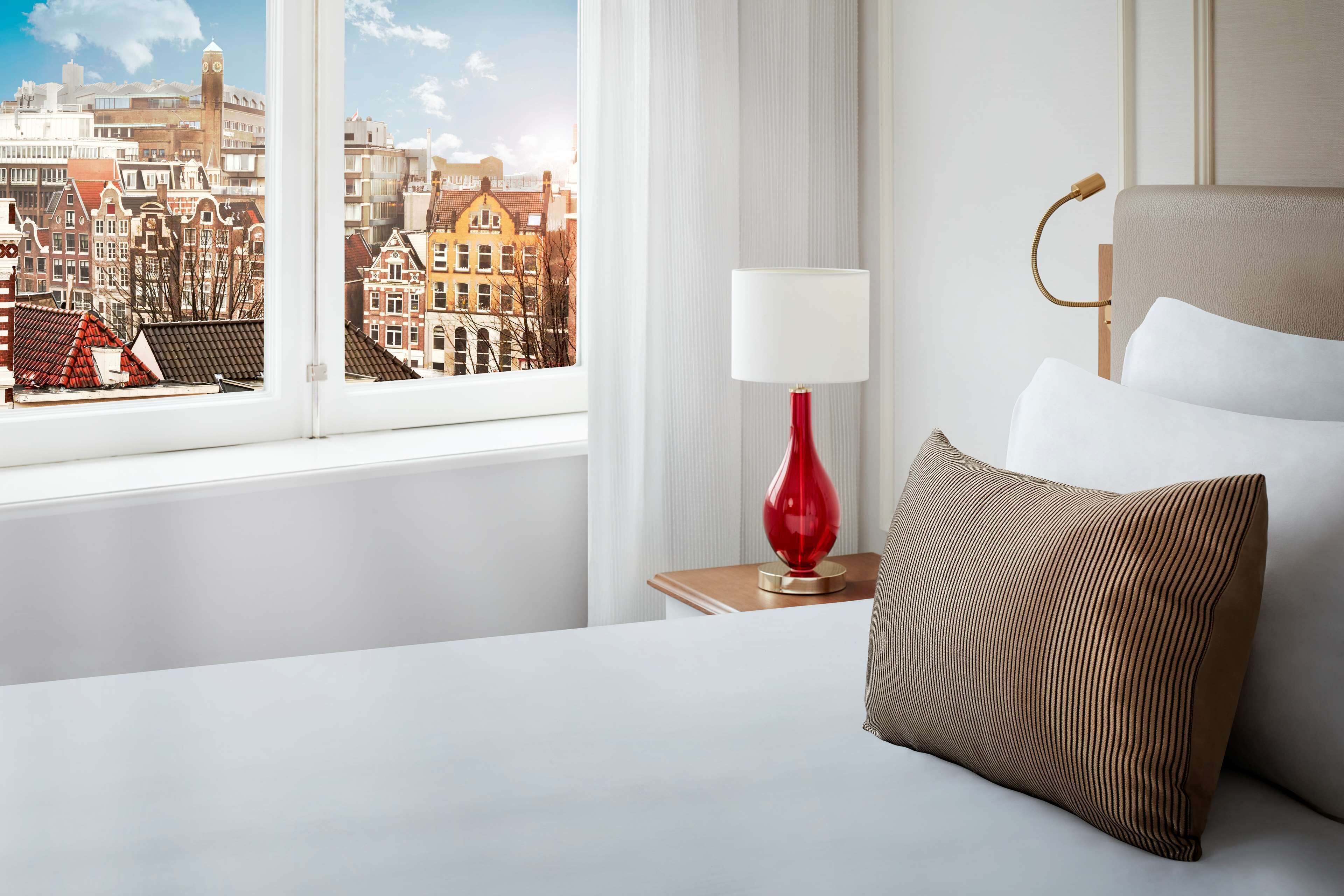 A hotel room with a bed, a nightstand with a lamp, and a view of a cityscape through a window.