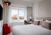 A hotel room with a large bed, a red chair, a desk with a lamp, and a view of a cityscape through the window.