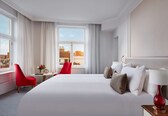 A hotel room with a large bed, a red chair, a small table with a vase, and a view of the city through large windows.