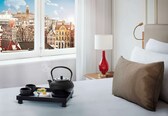 A hotel room with a bed, a nightstand with a lamp, a tray with tea set, and a window with a cityscape view.