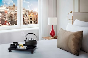 Et hotelværelse med en seng, et natbord med lampe, bakke med tesæt og et vindue med udsigt over bybilledet.