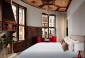 A hotel room with a large bed, a chandelier, a red chair, and a view of buildings through the window.