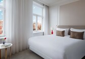 A hotel room with a large bed, a bedside table, a window with curtains, and a view of buildings.