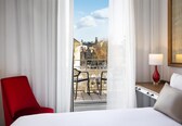 A hotel room with a bed, a red chair, a table with a lamp, and a view of buildings through a window.