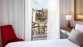 A hotel room with a bed, a red chair, a table with a lamp, and a view of buildings through a window.
