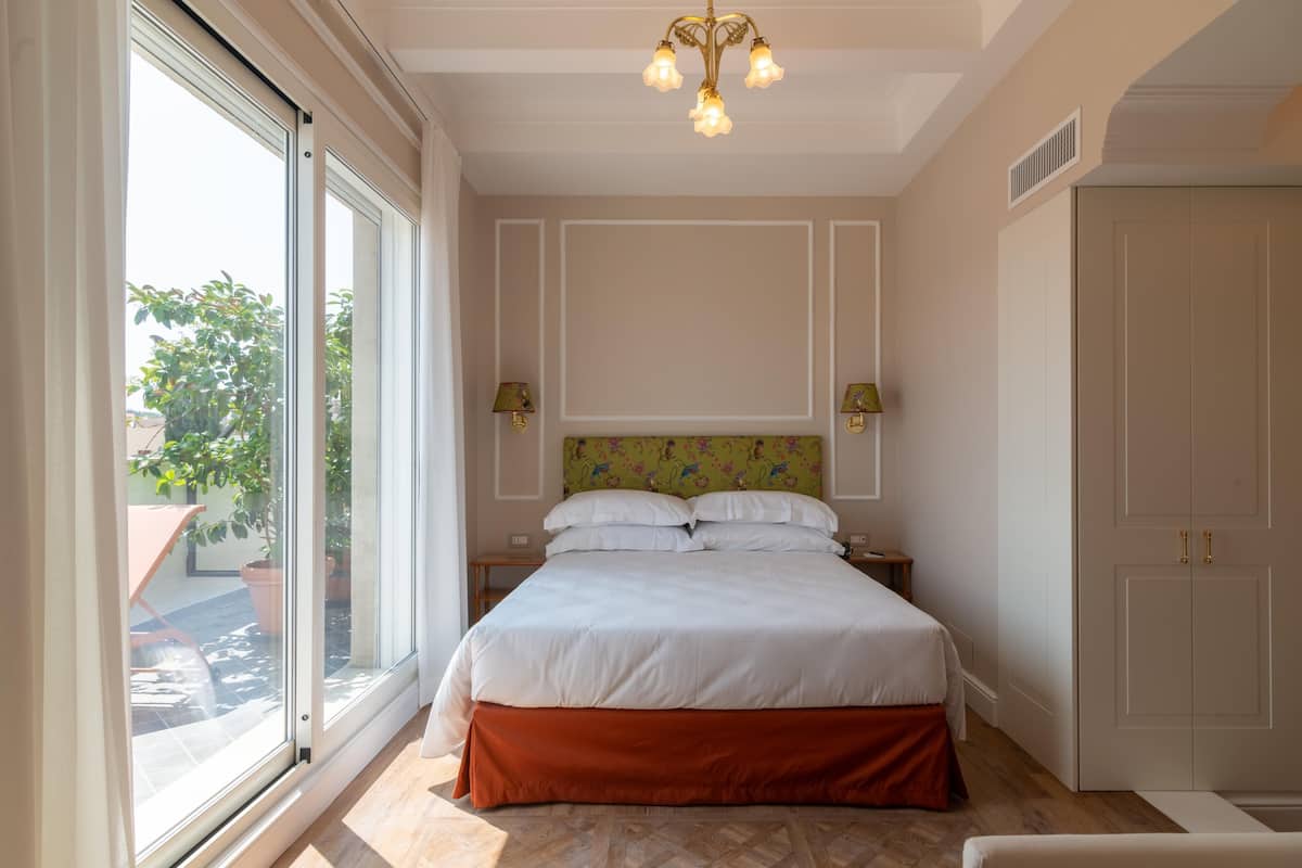 A bedroom with a large window, a bed with white bedding, two bedside lamps, and a chandelier.
