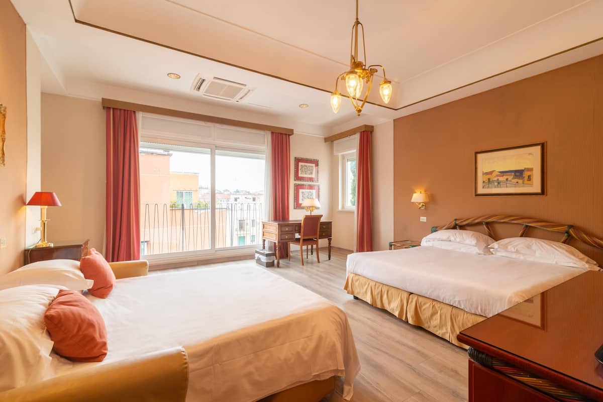 A hotel room with two beds, a desk, a chair, a balcony, and a chandelier.