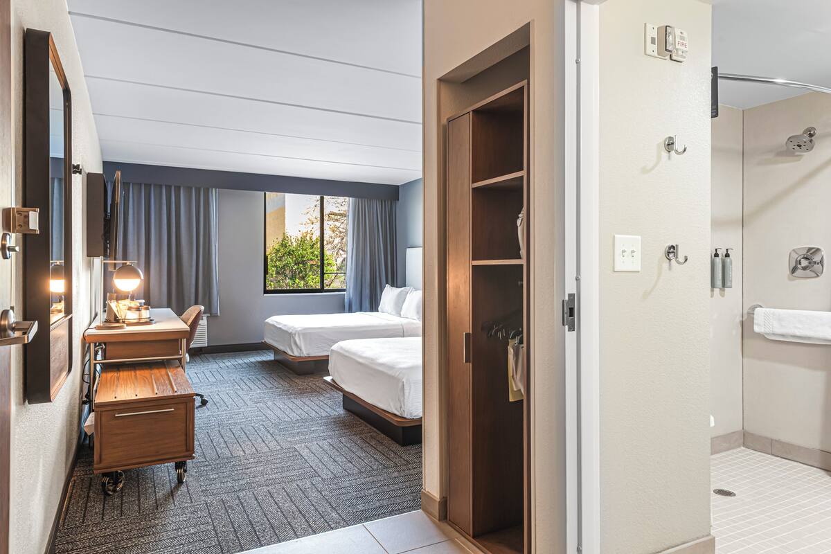 A hotel room with a desk, two beds, a wardrobe, and a shower area.